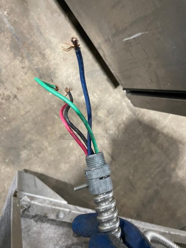 A close-up view of frayed electrical wires, including blue, green, red, and black, protruding from a metal conduit against a concrete floor.