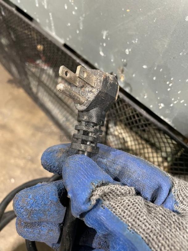 Close-up of a three-prong power plug held in a gloved hand. Plug appears well-used and slightly worn.