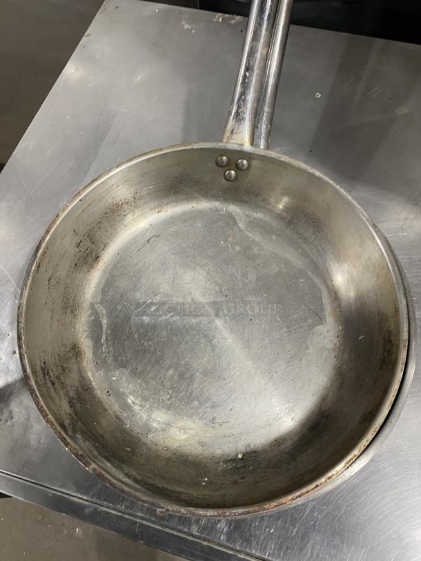 Commercial round frying pan with sturdy metal handle, shows signs of wear. Ideal for heavy-duty kitchen use.