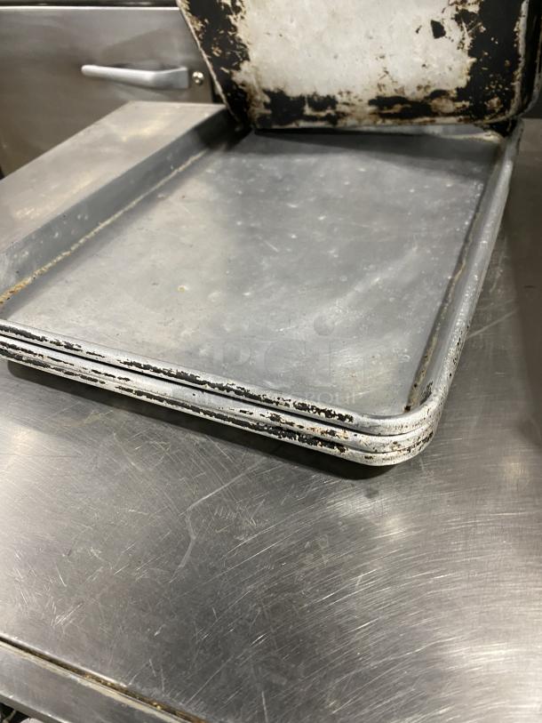 Commercial stainless steel full-size sheet pan, signs of heavy use, stacked, suitable for industrial kitchens or bakeries.