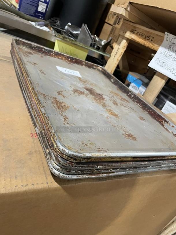 Commercial aluminum full-size sheet pans, stack of six, used condition with visible wear and patina, on a cardboard box.