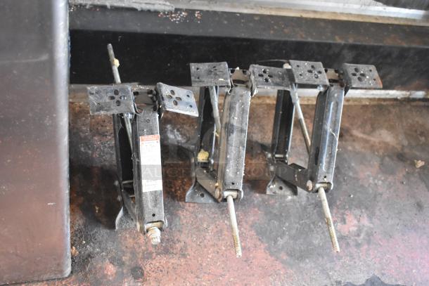 Four metal stabilizing jacks on a worn surface, showing usage wear. One jack has a warning label attached.