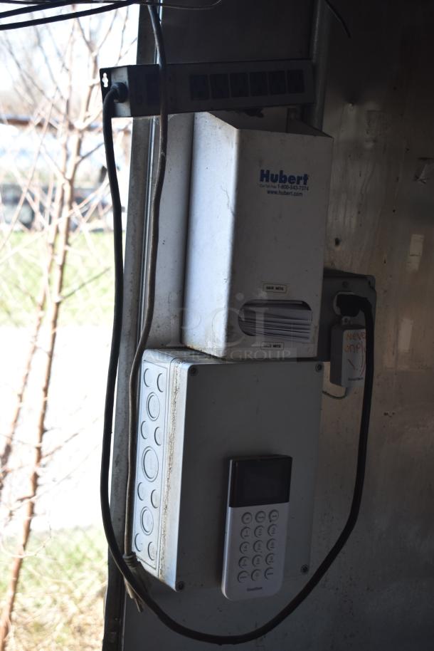Hubert hair net dispenser and Simplisafe keypad mounted inside a mobile kitchen trailer, with cords and outlets visible.