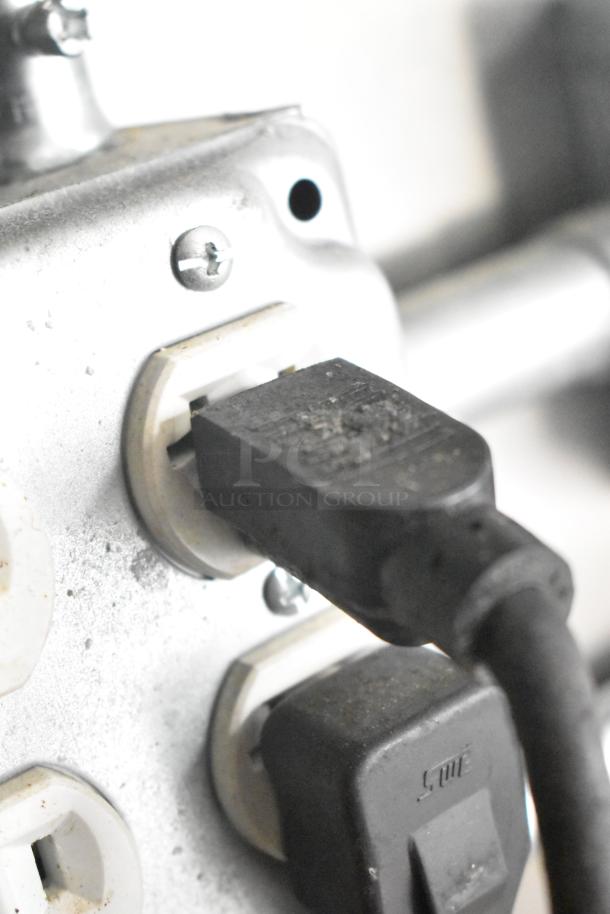 Power outlet with black plug connected, dust visible on the outlet surface, metallic frame with screws.