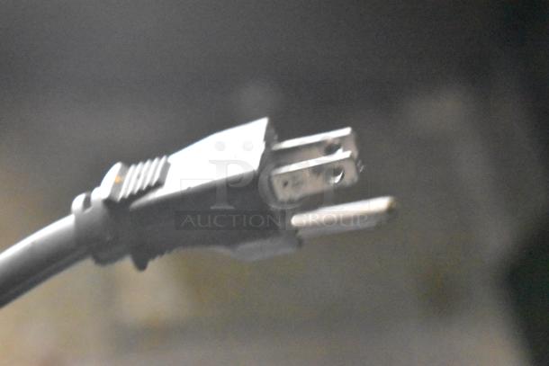 Close-up of an electrical plug with a three-prong design, attached to a black cable, against a blurred background.