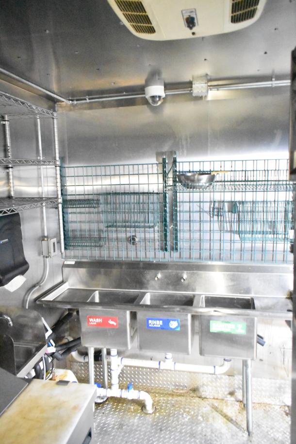 Interior of a 2021 Diamond Cargo 8.5x24 mobile kitchen trailer showing a triple sink setup, wire racks, and stainless steel finish.