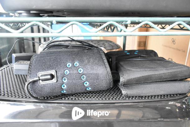 Four items including two LifePro vibrating plates, black neoprene sleeves with blue accents, and power cables on metal rack.