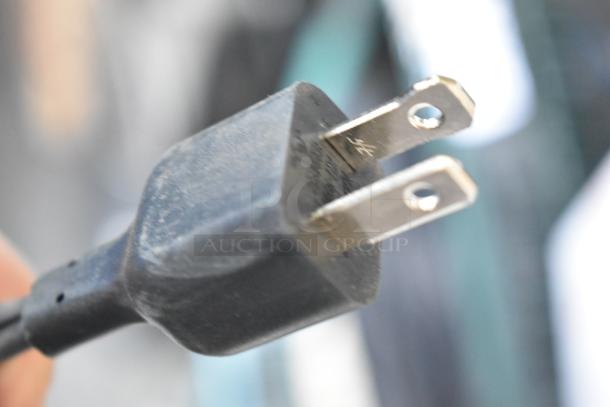 Close-up of a power plug, highlighting its prongs and casing. Part of an auction lot including Hurtle and LifePro vibration plates, Chefman pizza maker, and Harman Kardon speaker.