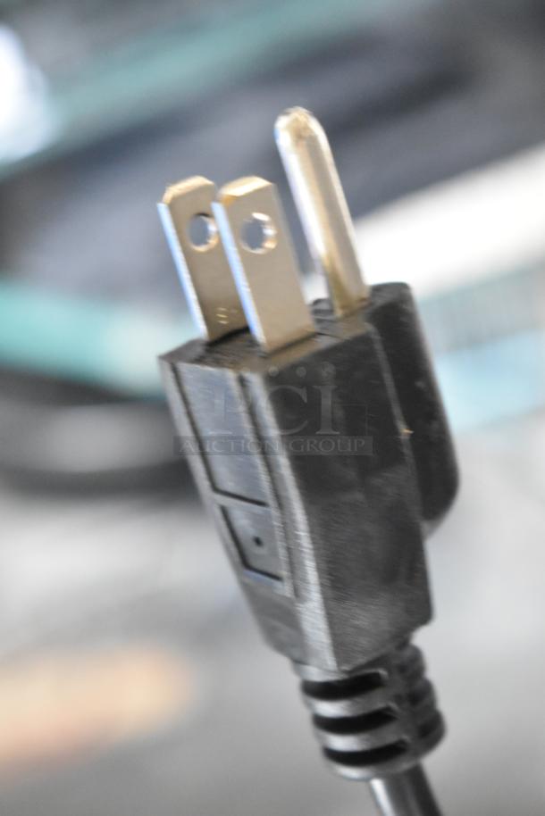 Close-up of a black three-prong power plug, part of the auction set including Hurtle HURVBTR36, LifePro plates, Chefman Pizza Maker, Harman Kardon Speaker.