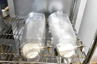 Two clear poly dry food dispenser hoppers on a metal shelf, each with a dispensing mechanism and a label indicating auction details.