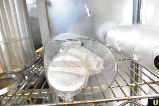 Two new clear poly dry food dispenser hoppers, oval-shaped, sealed in plastic, displayed on a metal rack.