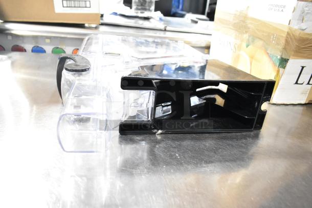 Clear poly dry dispenser hopper with black lid and handle, shown on a metal surface. Ideal for dispensing dry goods.