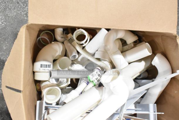 Box of various plumbing items including brackets, extension tubes, and hoses. Some items feature barcodes and labels.