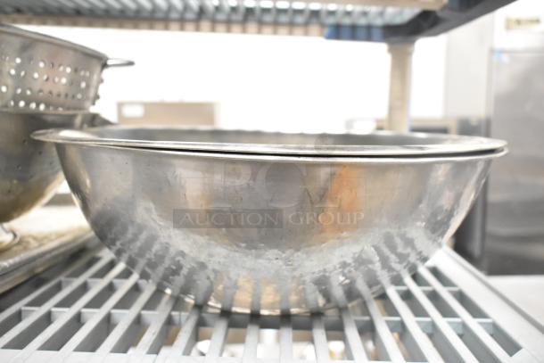 Stainless steel kitchen item assortment including bowls and colanders on metal shelf. Varied used condition.