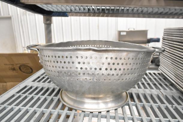 Metal colander on a shelf, perforations for draining, durable handles, part of a lot including bowls and baking pans.