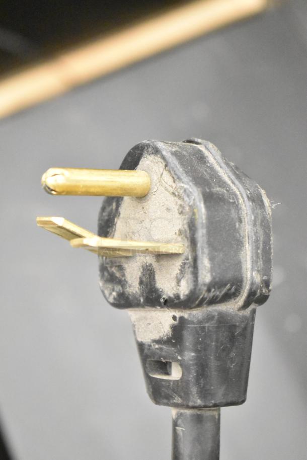 Close-up of a power plug for the Merrychef eikon e2s high-speed oven, showing three brass prongs on a black plastic body.
