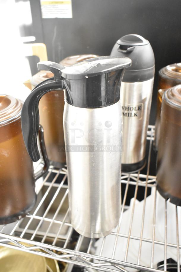 Stainless steel thermal pitcher with black plastic handle on a wire rack, next to brown and silver poly containers.