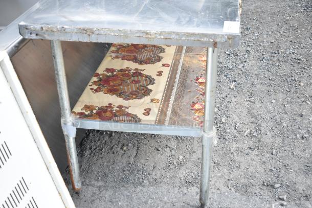 Commercial table with an under shelf, metallic finish, floral lining on shelf, shows signs of wear and rust.