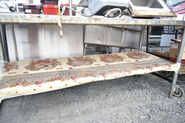 Commercial metal table with under shelf, showing signs of rust and wear. Decorated with a floral-patterned tablecloth.