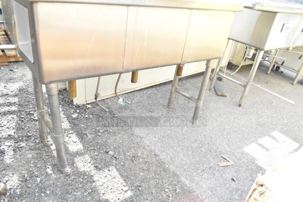 Commercial 3 bay stainless steel sink with sturdy legs, visible plumbing connections, and some surface wear. Ideal for kitchens.