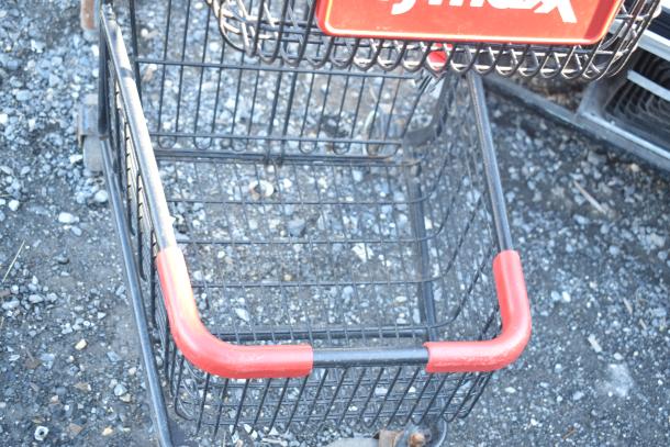 2 tier shopping cart with black metal frame and red handle. Shows a slightly worn condition. TJ Maxx sign attached.