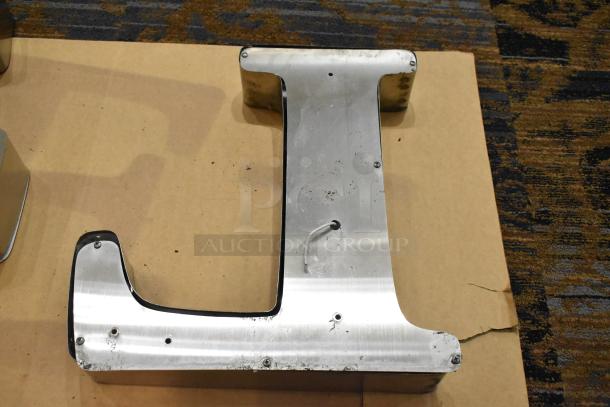 White "L" letter with a silver finish frame, visible wear and screw holes, lying on a cardboard surface.