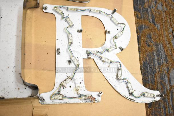 White letter "R" with silver finish frame, showing attached LED strip lighting on cardboard background.