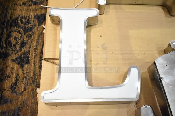 White letter "L" with silver finish frame, used condition, on cardboard surface, potential decorative signage.