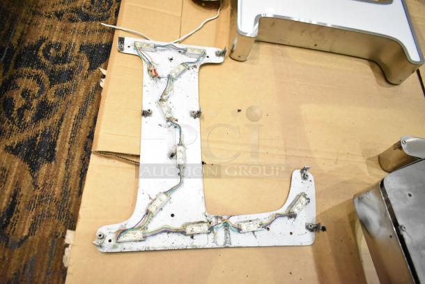 "White L letter with a silver finish frame, featuring internal wiring and mounting brackets, shown on cardboard surface."
