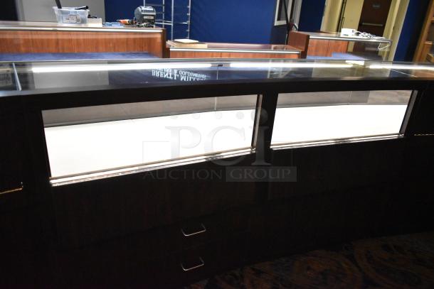 Lighted curved wooden jewelry display case with glass top, rear doors, and drawers, ideal for merchandising; buyer removal required.