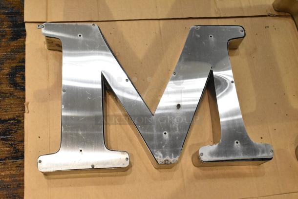 White "M" letter with a silver finish frame, metallic, visible screws, slight wear, approximately 2 feet tall.