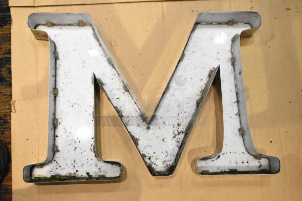 White letter "M" with silver finish frame, weathered condition.