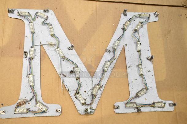 "White M letter sign with silver finish frame, featuring internal wiring and LED mounts; suitable for decorative use."