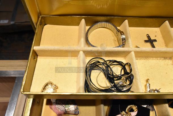 Gold jewelry case with assorted contents: silver watch, black cross pendant, gold heart pin, multiple necklaces.
