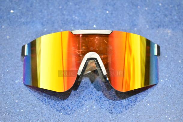 Brand new golden framed sunglasses with orange lens, reflecting a rainbow hue, set on a textured blue surface.