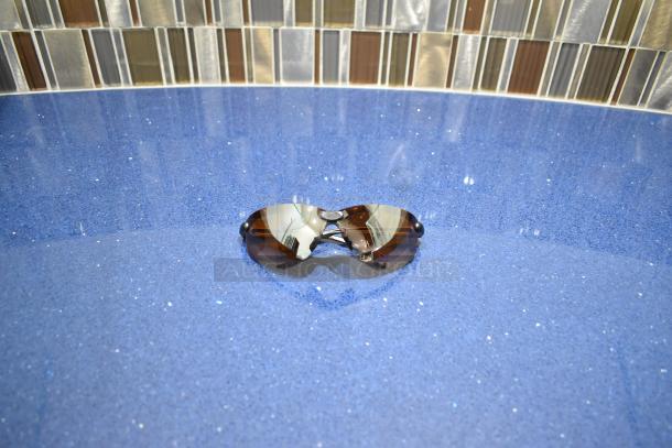 Brand new gray framed sunglasses with BluBlocker brown lens on a blue surface, reflecting a tiled background.
