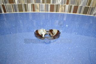 Brand new gray framed sunglasses with BluBlocker brown lens on a blue surface, reflecting a tiled background.