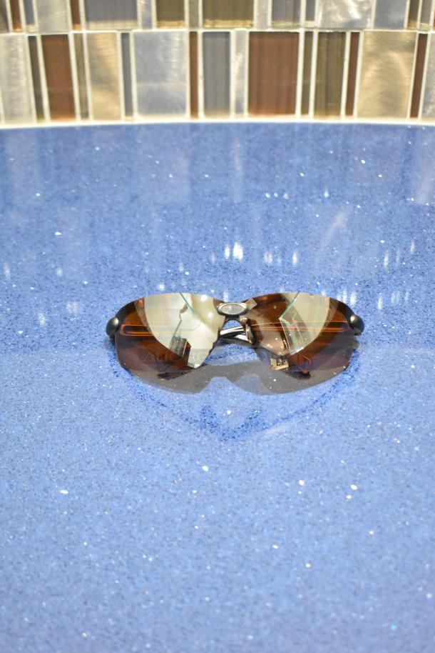 Brand new brown framed sunglasses with BluBlocker brown lenses, reflective finish, on a blue surface.