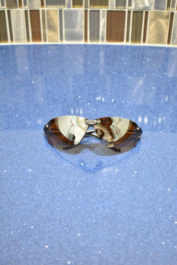 Brand new gray framed sunglasses with BluBlocker brown lenses, displayed on a blue surface.
