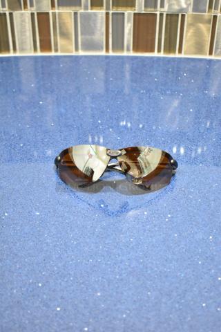 Brand new gray framed sunglasses with BluBlocker brown lenses, displayed on a blue surface.