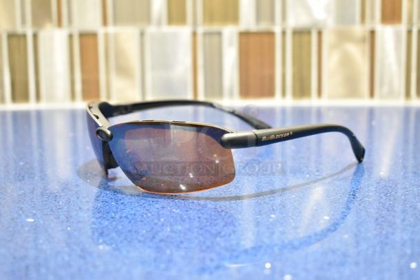 Brand new gray framed sunglasses with BluBlocker brown lenses on a blue surface, pristine condition.