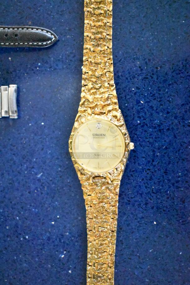 Gruen diamond quartz watch with textured gold-tone band, shown with black leather watch band and metal clasp, on blue surface.