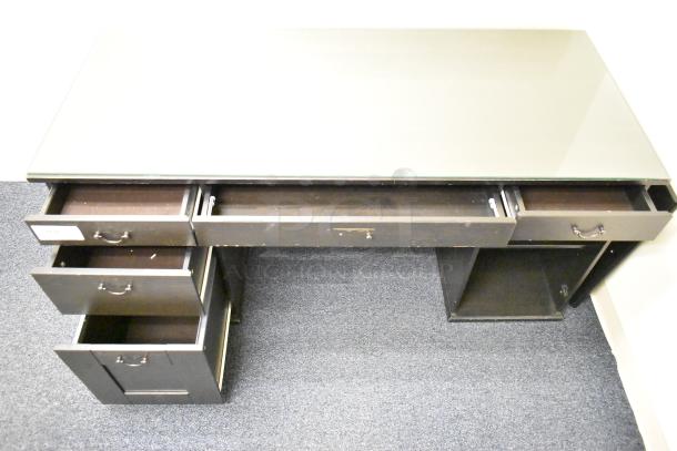 Wooden desk with five drawers, dark finish, glass top. Shows signs of use. Tag indicating auction number attached.