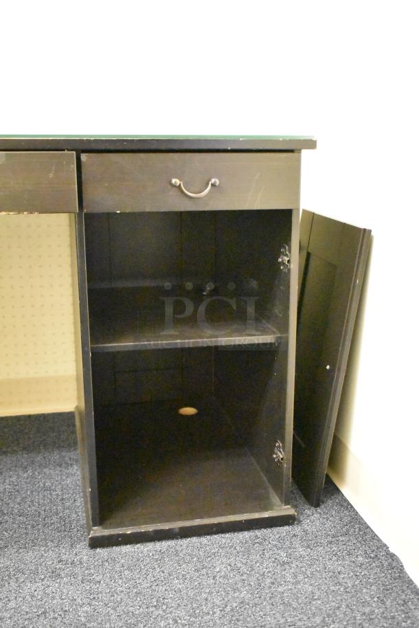 Wooden desk with five drawers, dark finish, and cabinet space. Features sturdy metal handles, showing moderate wear.
