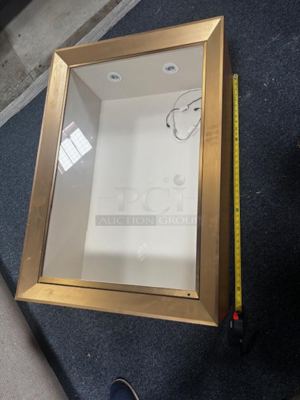 Gold-framed wall pop out display case with glass front; features internal lighting and visible wiring. Dimensions shown with tape measure.