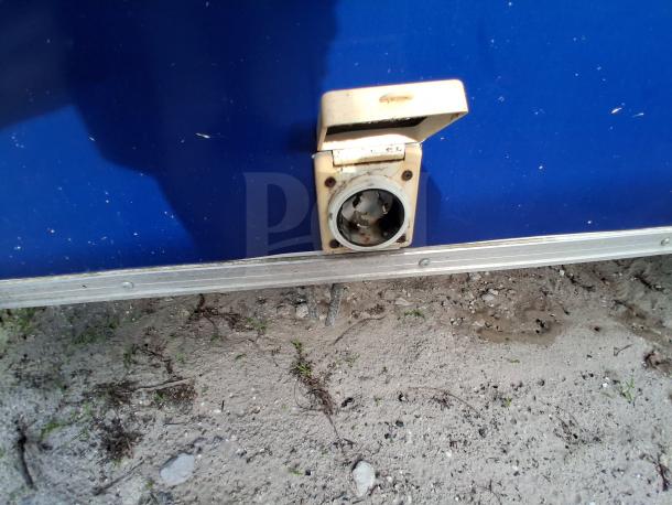 Food trailer exterior with exposed electrical outlet, blue paneling, and metal trim. Ground shows signs of outdoor placement.
