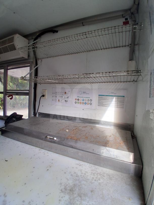 Food trailer interior with stainless steel countertop, wire shelving, wall instructions, and natural light through window.