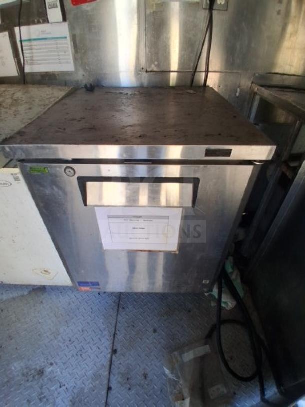 Stainless steel commercial refrigerator inside a food trailer. Features a visible label and utility connections. Some surface wear.