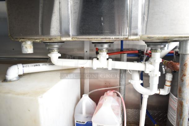 Under-sink piping and containers in a commercial mobile kitchen trailer. Features Ecolab cleaning supplies.