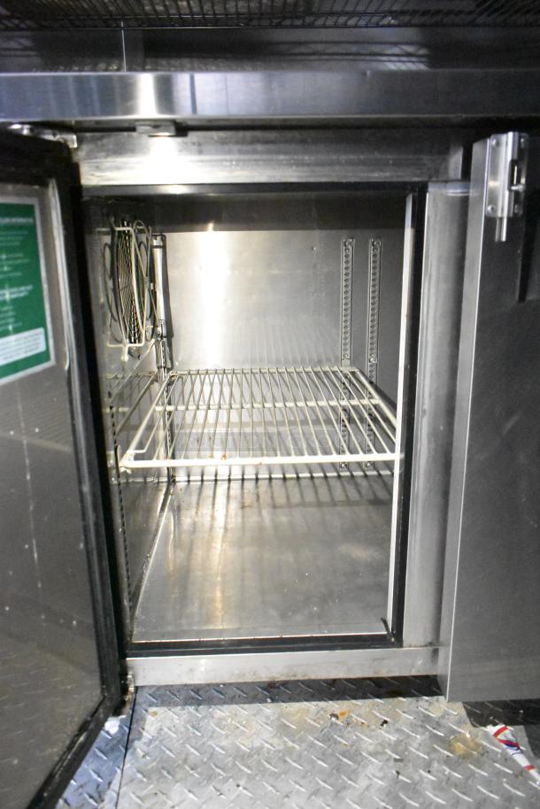 Interior of commercial mobile kitchen trailer, featuring stainless steel refrigeration unit with metal shelving and wiring.